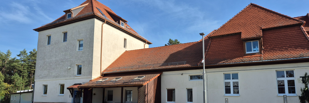 Blick auf das Dienstgebäude der Geschäftsstelle der AG Flussgebietsbewirtschaftung. Zu sehen ist ein dreigeteiltes gelbes Gebäude mit rotem Dach vor blauem Himmel. Der linke Teil des Gebäudes ist höher als der Rest.